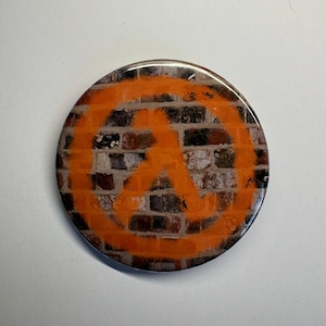 May include: A circular badge featuring an orange symbol set against a brick wall design. The symbol is a stylized logo. The badge has a shiny surface.
