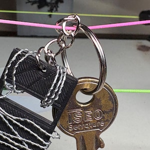 May include: Close-up of a key ring with keys and a black tag. The key ring is silver and holds a brass key with the text "ISEO Serrature". The black tag is wrapped with silver wire. A pink string is visible in the background.