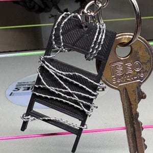 May include: A black miniature chair keychain with silver wire detailing. The keychain is attached to a silver keyring and a gold-colored key with the text "ISEO Serrature". The background is blurred with pink and green lines.