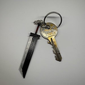 Peut inclure: Un porte-clés avec un anneau en métal argenté tenant une clé dorée et un porte-clés en forme d'épée noire et argentée. Le porte-clés a une poignée rouge et une lame détaillée.