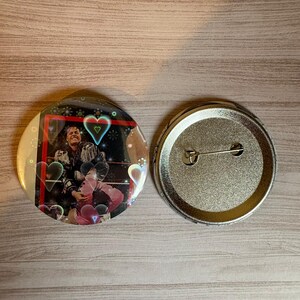 Könnte beinhalten: Ein runder Ansteckbutton mit holografischer Beschichtung und Herzmotiven. Die Vorderseite zeigt ein Foto einer Person in dunkler Jacke und rosa Hose, umrahmt von einem roten Rand. Die Rückseite ist aus silberfarbenem Metall mit einer Nadel.