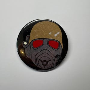 May include: A round button featuring a stylized illustration of a helmet with red lenses and a respirator. The helmet is tan, and the respirator is gray. The button has a black border and is set against a white background.