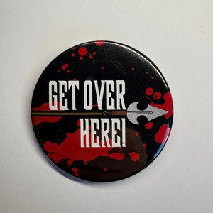 Peut inclure: Bouton rond noir avec le texte "GET OVER HERE!" en lettres blanches. Le bouton présente un graphique de lance et des éclaboussures rouges. Un article de fantaisie.