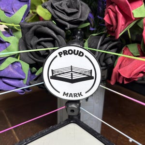 May include: A decorative arrangement featuring a wrestling ring emblem with the words "PROUD MARK" on a white circular background. The arrangement includes black, purple, and red artificial roses.