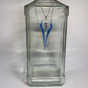 Può includere: Collana color argento con un ciondolo dal design blu e bianco. Il ciondolo ha una forma stilizzata con un piccolo accento viola. La collana è esposta all'interno di un barattolo di vetro trasparente.