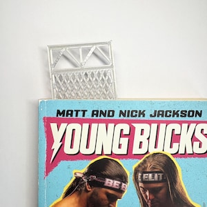 May include: A silver metal decorative object, possibly a bookmark, rests atop a book titled "Young Bucks." The book cover features illustrations of two men wearing headbands. The text "Matt and Nick Jackson" is above the title.