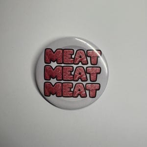 Puede incluir: Un botón redondo blanco con la palabra "MEAT" apilada tres veces en una fuente roja audaz. El botón tiene un acabado brillante y un diseño simple y limpio, perfecto para añadir un toque de humor a la ropa o accesorios.