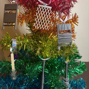 May include: A rainbow-colored tinsel Christmas tree decorated with miniature ornaments. The ornaments include a miniature chair, a chain-link fence, a magnifying glass, and a small wooden object. The tree is festive and colorful.