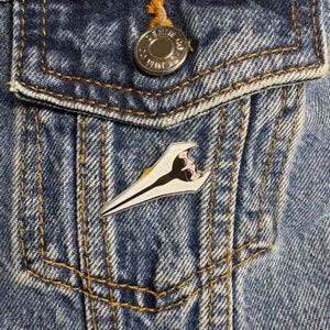 May include: A triangular enamel pin featuring a stylized illustration of a figure with long black hair and a pale face, set against a blue denim jacket with a silver metal button embossed with the text Denim Co.