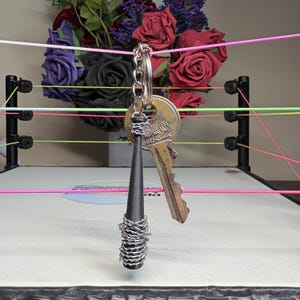 Puede incluir: Un llavero con una llave dorada y un dije negro en forma de cono envuelto con alambre plateado. La llave tiene el texto "SEO Serretture". El fondo incluye un arreglo floral y un ring de lucha libre.