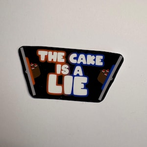 May include: A black sticker with the text "THE CAKE IS A LIE" in white, orange, and blue. The sticker has a rectangular shape with angled sides and features cartoon cake illustrations.