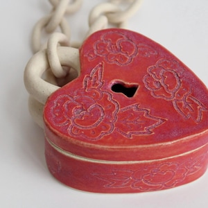 May include: A heart-shaped ceramic box in a vibrant red glaze with etched floral patterns and a central keyhole cutout. The box is attached to a thick, off-white ceramic chain with large, rounded links that extend from the top of the heart.