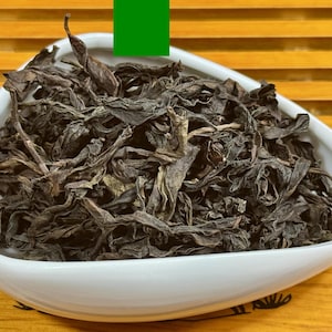 May include: A white, oval-shaped bowl filled with dried, dark brown tea leaves. The tea leaves have a wrinkled texture and are piled high in the bowl. The bowl sits on a wooden surface with a slatted design.