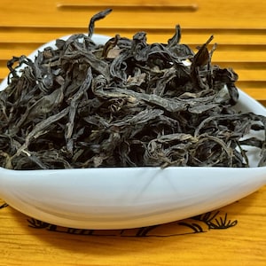Puede incluir: Un cuenco blanco de forma ovalada lleno de hojas de té oscuras y secas. Las hojas tienen un color marrón oscuro y una apariencia texturizada. El cuenco está sobre una superficie de madera con un tono amarillo.