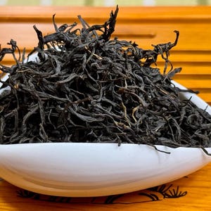 Puede incluir: Primer plano de té negro en hojas sueltas en un cuenco de cerámica blanco. Las hojas de té son de color marrón oscuro y tienen una forma ligeramente rizada.