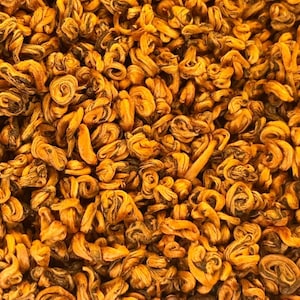 May include: Close-up of a pile of dried tea leaves. The leaves are a mix of golden and brown hues, with a twisted, curled appearance. The texture appears dry and slightly coarse, suggesting a high-quality tea product.