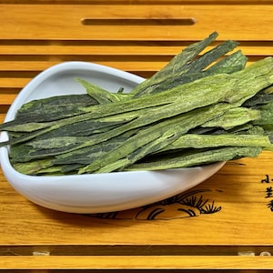 May include: A white, oval-shaped bowl filled with dried green tea leaves. The tea leaves are long and flat, with a slightly curled appearance. The bowl sits on a wooden tray with a slatted design.