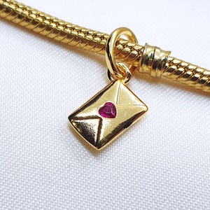 May include: A gold-colored charm featuring an envelope design with a small, heart-shaped, red gemstone. The charm is suspended from a gold-colored, snake chain bracelet. The background is a white, textured fabric.