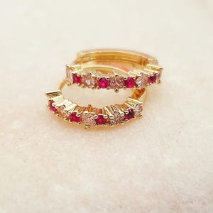Può includere: Due orecchini a cerchio color oro con una fila di pietre preziose rosse e chiare alternate. Gli orecchini sono esposti su una superficie bianca strutturata. Le pietre preziose sono piccole e ben distanziate.