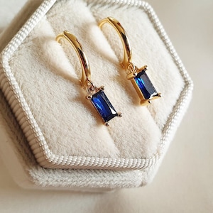 May include: A pair of gold-colored hoop earrings with rectangular, deep blue gemstones. The earrings are displayed in a light gray velvet jewelry box with a hexagonal shape. The background is a soft, neutral color.