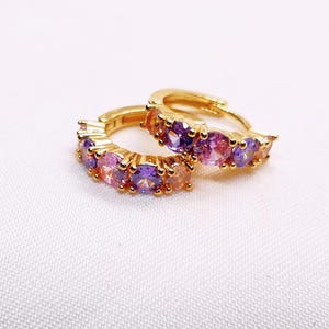 Puede incluir: Un par de pendientes de aro dorados con una fila de piedras preciosas de colores púrpura, rosa y melocotón. Los pendientes están sobre un fondo blanco.