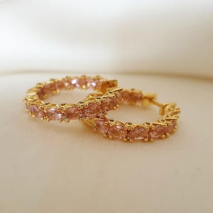 May include: A pair of gold-colored hoop earrings, each encrusted with small, light pink gemstones. The earrings are displayed on a soft, off-white surface, creating a contrast with the jewelry's warm tones. The gemstones are evenly spaced around the hoops.