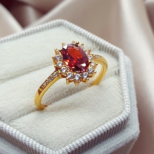 May include: A gold ring featuring a large oval, red gemstone at its centre, encircled by smaller white gemstones. The ring's band is also decorated with small white gemstones. The ring is presented in a grey velvet jewellery box.