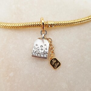 Könnte beinhalten: Ein goldfarbenes Armband mit einem silbernen Glockenanhänger, der mit Kristallen besetzt ist. Ein kleiner goldener Anhänger mit der Aufschrift "Tea Time" hängt daran. Das Armband liegt auf einer weißen Oberfläche.