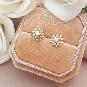 May include: A pair of gold stud earrings shaped like multi-layered flowers, each featuring a central clear round gemstone. The earrings are displayed in a beige velvet hexagonal jewelry box, positioned between two white artificial roses.