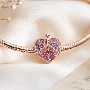 Può includere: Un bracciale a catena snake color oro rosa con un ciondolo a forma di foglia. Il ciondolo è tempestato di piccoli cristalli scintillanti rosa e viola disposti a sfumatura, che mettono in risalto le venature dettagliate della foglia.