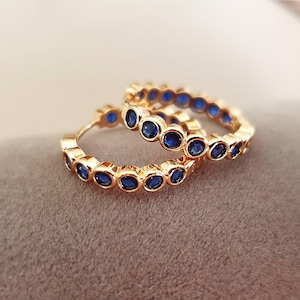 Könnte beinhalten: Ein Paar goldfarbene Creolen. Jeder Ohrring ist mit einer Reihe kleiner, oval geformter, tiefblauer Edelsteine verziert. Die Ohrringe werden auf einer weichen, neutral getönten Oberfläche präsentiert.