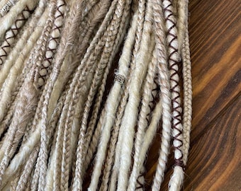 May include: A close-up of a bundle of synthetic dreadlocks in various shades of beige and white. Some of the dreadlocks have brown leather stitching and silver beads.