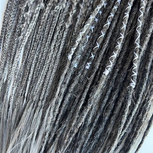 May include: Close-up of gray and white dreadlocks. The dreadlocks are a mix of gray and white, with some sections featuring white thread accents. The texture of the dreadlocks is visible, with a combination of smooth and textured strands.
