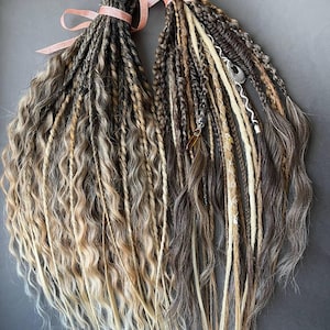 Peut inclure: Deux ensembles de dreadlocks dans des tons de marron, blond et gris, attachés avec des rubans roses. Les dreadlocks présentent un mélange de textures, avec des sections ondulées et droites, sur fond gris.