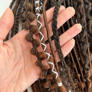 May include: A close-up of a hand holding a bundle of brown and black synthetic dreadlocks with white stitching. The dreadlocks are arranged in a fan shape, with the stitching visible on the top of the dreadlocks.