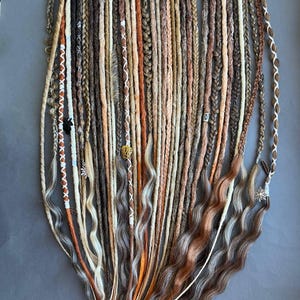 May include: A selection of handmade dreadlocks in shades of brown, blonde, and orange. Some are braided, others wavy, and some are decorated with beads and charms. The dreadlocks are displayed on a grey surface.