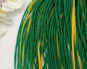 May include: A bundle of green and yellow dreadlocks, tied together at the top with twine. The dreadlocks are a vibrant green with strands of bright yellow woven throughout. The background is white, with a white orchid flower and brown buds in the upper left corner.
