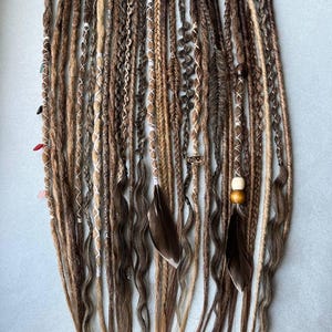 May include: A collection of handmade dreadlocks in various shades of brown and blonde. Some dreads are adorned with white thread, beads, and feathers. The dreadlocks are arranged against a light blue background, showcasing their diverse textures and styles.