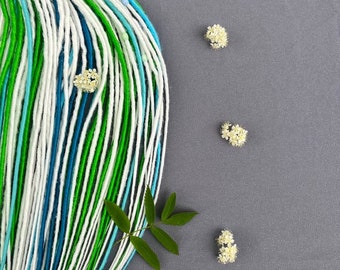 May include: A bundle of synthetic dreadlocks in white, green, and blue colors. The dreadlocks are arranged in a fan shape and are lying on a gray surface. There are small white flowers scattered around the dreadlocks.