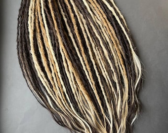 May include: A bundle of handmade dreadlocks in various shades of brown, blonde, and white. The dreads are arranged in a neat, oval shape, with a mix of dark and light colors creating a natural, textured appearance. The ends are slightly frayed.