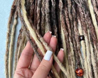 May include: Close-up of handmade dreadlocks in various shades of brown, beige, and blonde. The dreads are textured and appear to be made of synthetic or natural hair. A few strands are held by a hand with white nail polish. Decorative beads are attached.