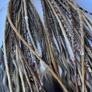 May include: A collection of handmade dreadlocks in various shades of brown, beige, and gray. Some dreads are adorned with decorative wraps, beads, and charms. A dark feather is also included in the arrangement, adding a natural element to the design.