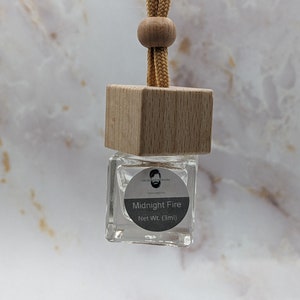 May include: A small, clear glass bottle with a wooden cap and a yellow string hanging from it. The bottle has a label that says "Midnight Fire" and "Net Wt. (3ml)".