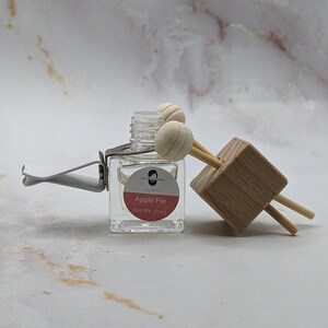 May include: A small glass bottle with a clear liquid and a white label that says "Apple Pie" and "Net Wt. (3ml)". The bottle is attached to a wooden car air freshener with two white diffuser balls.