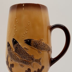 May include: A brown ceramic mug with a light brown glaze features three fish in relief. The fish are brown and have a textured surface. The mug has a brown handle.