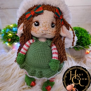 以下が含まれることがあります： 緑の白と赤のストライプのドレスを着た、白いサンタの帽子をかぶった、かぎ針編みのエルフのドール。ドールは茶色の髪と茶色の目です。