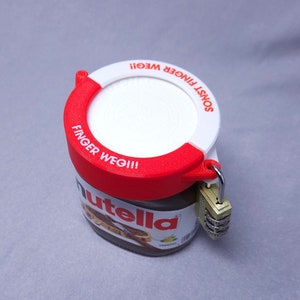 Könnte beinhalten: Ein roter und weißer Plastikdeckel mit dem Text "SONST FINGER WEG!!" und "FINGER WEG!!" darauf. Der Deckel ist an einem Glasgefäß mit Nutella befestigt, das mit einem Zahlenschloss versehen ist.