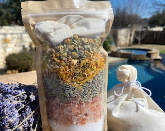 May include: A clear plastic bag filled with a layered bath soak. The bath soak includes dried lavender, chamomile, and calendula flowers, pink Himalayan salt, and white Epsom salt. The bag is sitting on a wooden surface with a white muslin bag next to it.