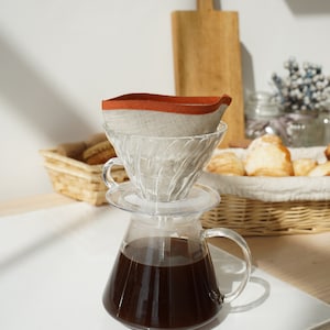 Op de afbeelding: Een glazen koffiekan met een helder glazen pour-over filter en een linnen koffiefilter met een rode rand. De kan is gevuld met donkere koffie en staat op een wit oppervlak.