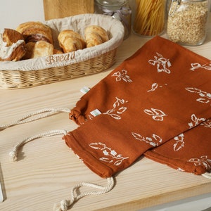 May include: Two brown reusable produce bags with white floral patterns and white drawstrings. The bags are lying on a wooden surface with a wicker basket of bread rolls in the background.
